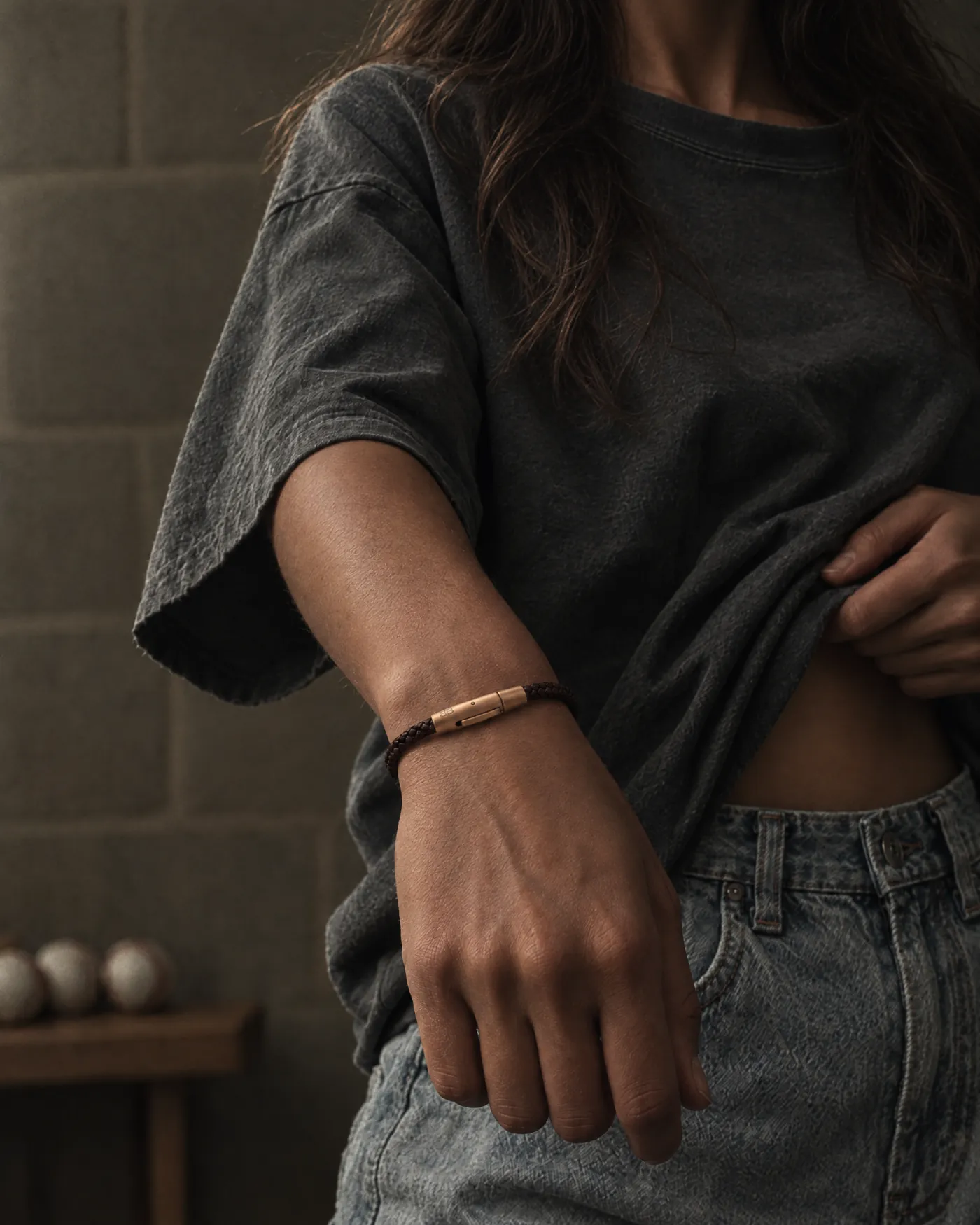 Thin brown braided leather bracelet with a rose-gold clasp, worn on a woman's wrist in front of a concrete wall as she lifts her t-shirt at the waist