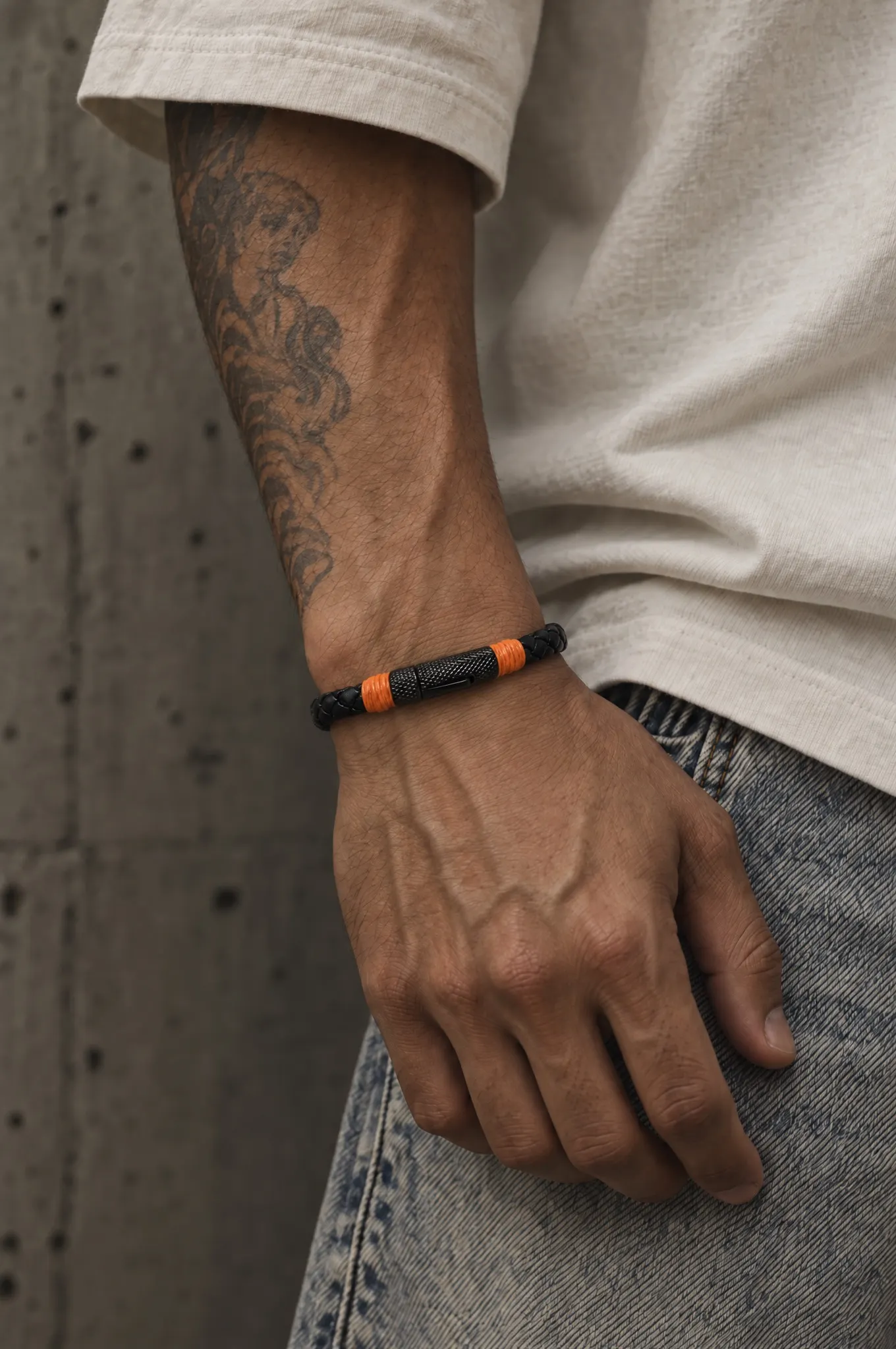 Black braided leather bracelet with orange wraps and a black clasp, worn on a tattooed forearm against a concrete wall