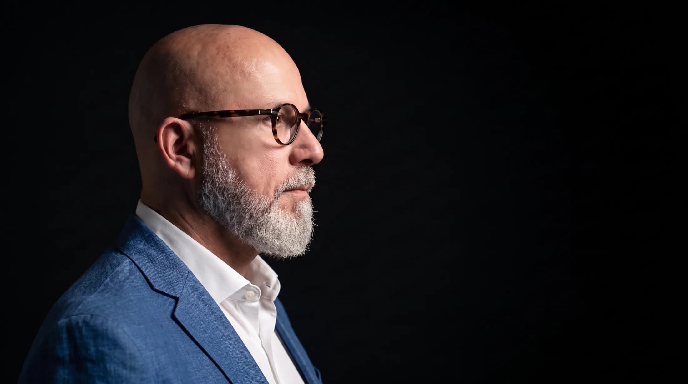 Daniel Valsjö in profile against a black background, wearing a blue linen jacket over a white shirt and tortoiseshell glasses